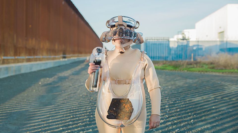 Artist Tanya Aguiñiga wearing a suit made of glass elements while standing next to the U.S./Mexico border wall