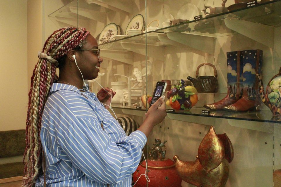 A woman stands in front of a case filled with sculptures and points her phone at them