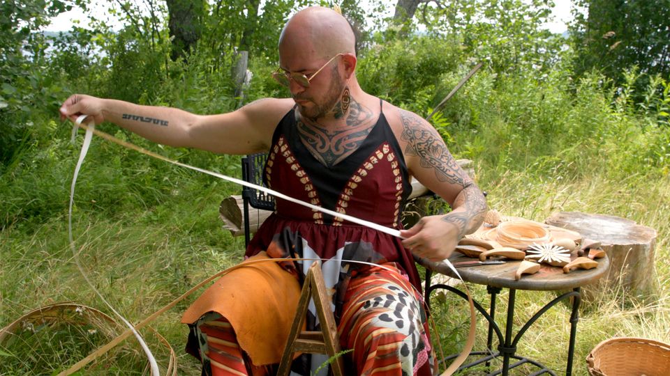 A bald man whose arms are covered in tattoos sits outside. He is holding canes for basketry.
