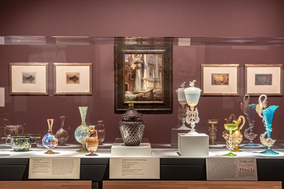 Exhibition gallery showing cases of glass artworks in the foreground and paintings against a dark red wall in the background.